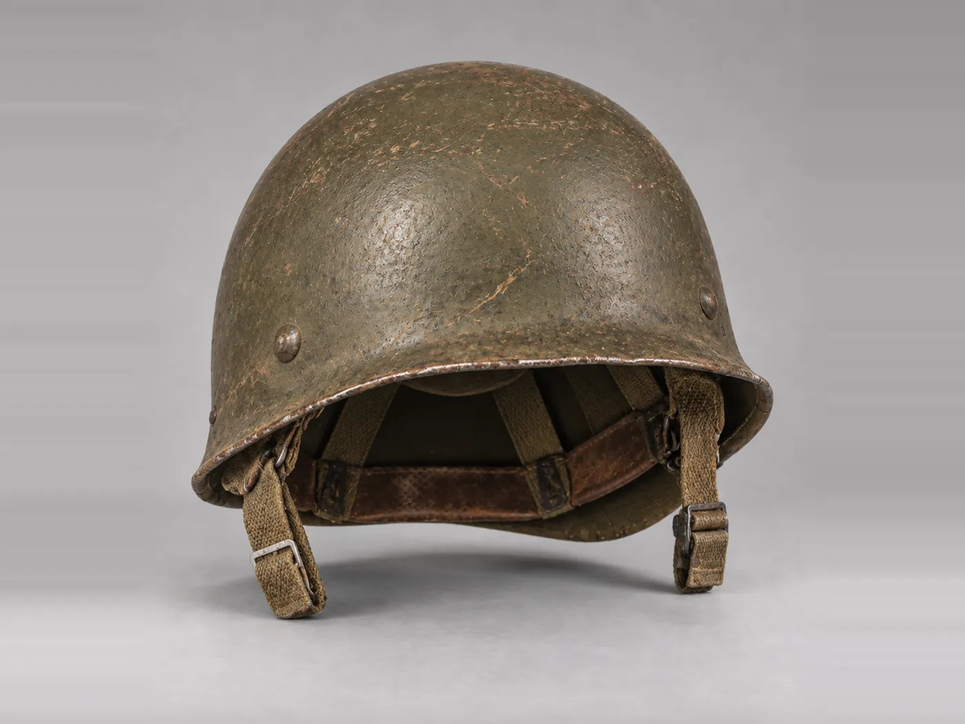 A worn olive drab steel helmet with a leather sweatband and canvas chin straps, photographed on grey